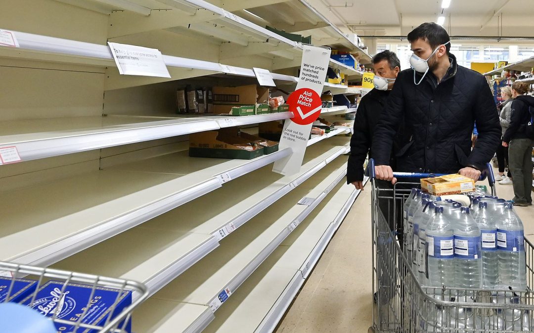 Customers in facemasks look at empty shelves in a grocery store.
